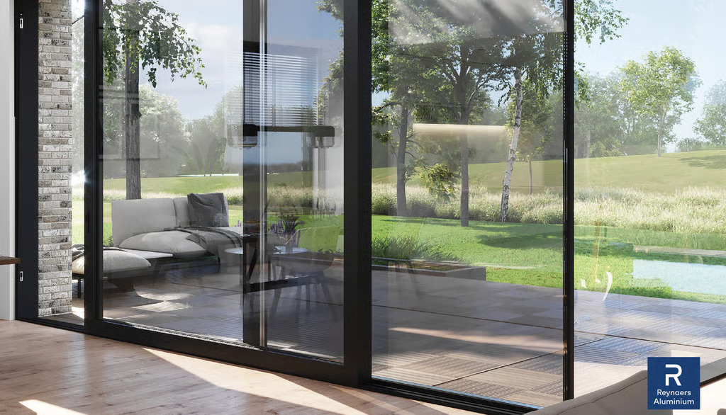 View from inside a modern living room looking through large black-framed sliding glass doors onto a sunny patio and green lawn.